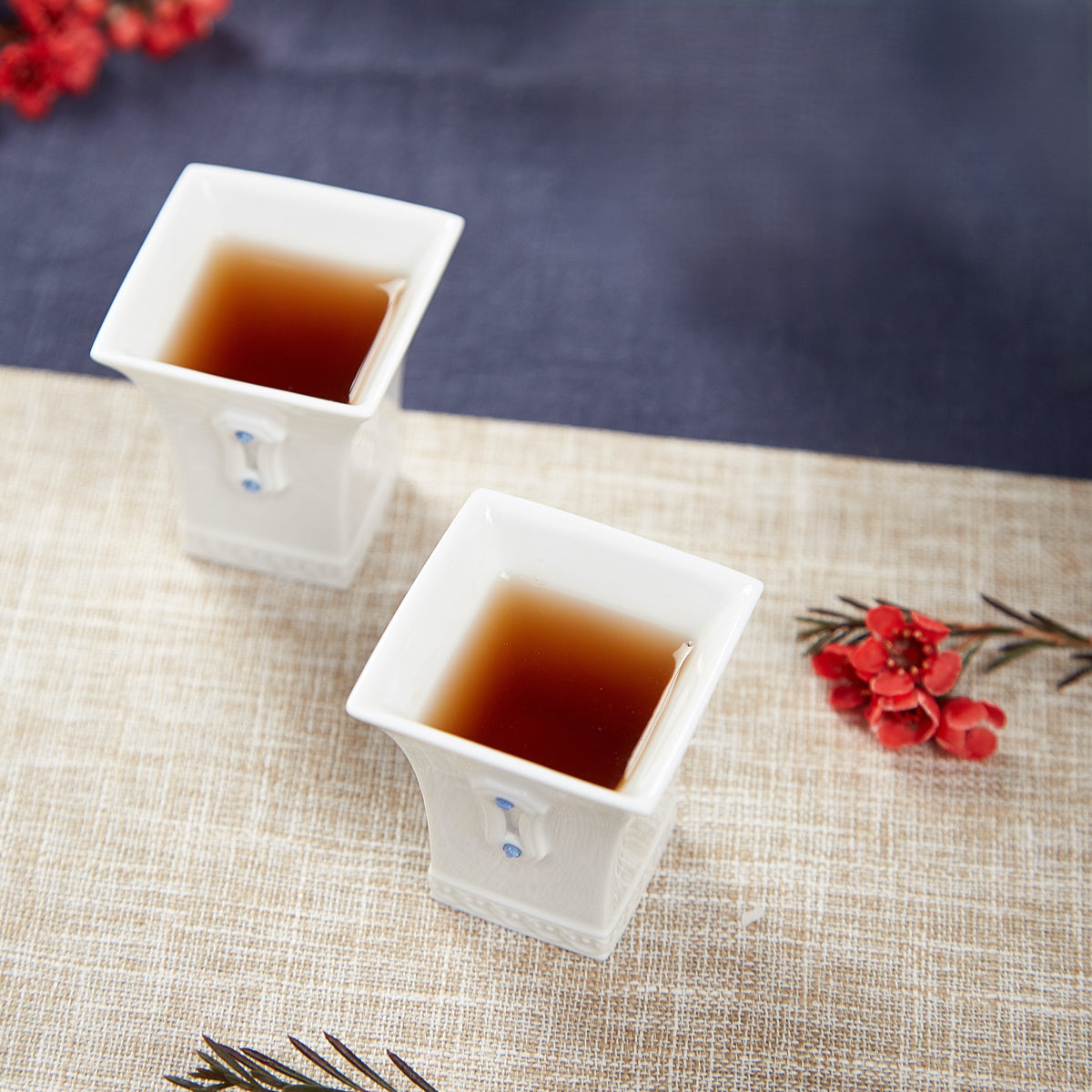 明珠在握兩只白瓷茶杯盛裝茶湯，方形杯口與藍色水鑽細節置於米色茶席